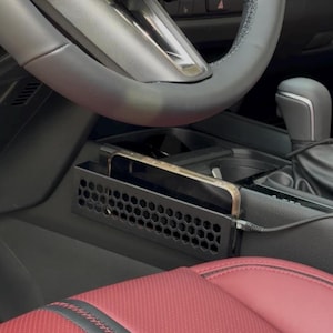 May include: A black metal phone holder with a honeycomb pattern is installed in a car's console. A smartphone is placed inside the holder and is connected to a charging cable. The car's steering wheel and red leather seat are visible.