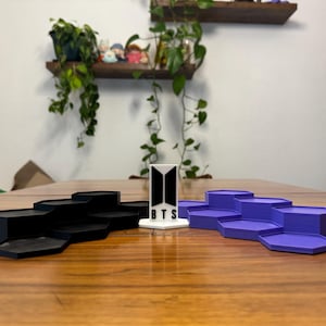 May include: Three-piece display stands in black and purple, with a white stand featuring the BTS logo and text. The stands are hexagonal and tiered, suitable for displaying collectibles or small items.