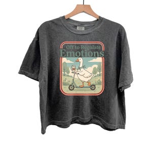 Può includere: T-shirt grigio scuro con una grafica di un'oca su uno scooter, che tiene una tazza di caffè, e la scritta "Off to Regulate Emotions". Il design è in stile retrò. La maglietta è appesa a una gruccia di legno.