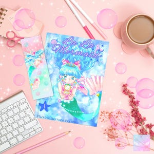 Puede incluir: Ilustraci&oacute;n colorida de una sirena en la portada de un libro y un marcap&aacute;ginas, con el t&iacute;tulo "La La Mermaid". La sirena tiene el pelo azul y la cola verde. Burbujas rosas y art&iacute;culos de papeler&iacute;a rodean el libro sobre una superficie rosa.