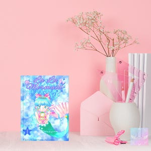 Puede incluir: Un libro titulado "La La Mermaid" con una ilustraci&oacute;n de sirena de dibujos animados. La portada presenta una sirena de pelo azul sosteniendo una perla. El libro se muestra con papeler&iacute;a rosa, un jarr&oacute;n de flores y un l&aacute;piz blanco.