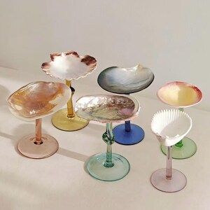 May include: A collection of six decorative glass pedestals featuring irregular, shell-shaped bowls in various colours including white, amber, blue, and pink, each supported by a slender stem and a circular base in coordinating or contrasting glass hues.