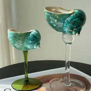 May include: Two decorative wine glasses with teal seashell bowls and clear stems. One glass has a green base and stem, the other has a clear base and stem. The glasses are on a white tray with the word "CELINE" printed on it.