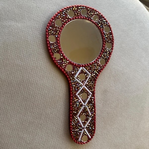 May include: A hand mirror with a round reflective surface, surrounded by a red border and small, round mirrors. The handle features a decorative pattern of white and gold accents, creating a unique and ornate design.