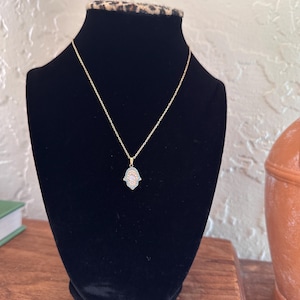 Könnte beinhalten: Eine goldfarbene Halskette mit einem Hamsa-Hand-Anhänger, präsentiert auf einer schwarzen Samtbüste. Der Anhänger hat ein detailliertes Design mit hellblauen und weißen Akzenten. Die Halskette wird auf einer Holzoberfläche präsentiert.