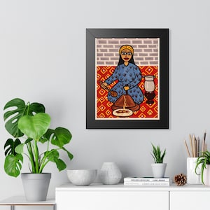 May include: Framed art print depicting a woman in a blue dress cooking over a fire. The artwork features a red and yellow patterned rug, a brick wall background, and a white pitcher. The woman is wearing a yellow headband.