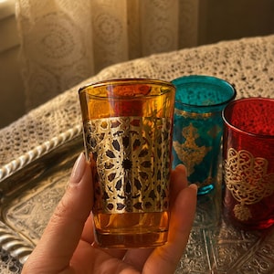 Op de afbeelding: Een amberkleurig glas met een gouden filigraanpatroon wordt in een hand gehouden. Twee andere glazen, een turquoise en een rode, zijn zichtbaar. De glazen staan op een zilveren dienblad, met een kanten gordijn op de achtergrond.