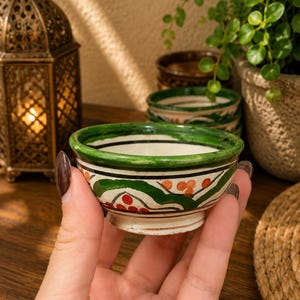 May include: A small, hand-painted ceramic bowl with a white base, green rim, and decorative floral patterns in red and orange. The bowl is held in a hand, with other similar bowls and a decorative lantern in the background. The bowl is approximately 2 inches in diameter.