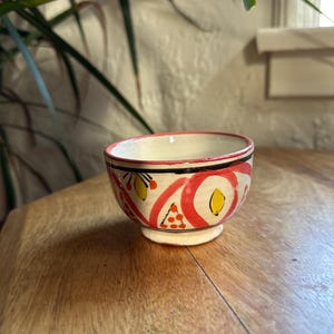May include: A small, hand-painted ceramic bowl with a white interior and a decorative exterior. The bowl features a red, black, and yellow design. The bowl is sitting on a wooden surface.
