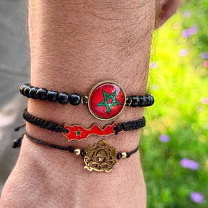 Può includere: Tre braccialetti con disegni marocchini. Uno ha perline nere e un emblema rosso e verde. Un altro ha un contorno rosso del Marocco con una stella verde. Il terzo braccialetto ha un emblema dorato.