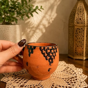 May include: A small, orange ceramic cup with a handle, decorated with black and green abstract patterns. The cup sits on a beige, crocheted doily. A decorative lantern and potted plant are in the background.