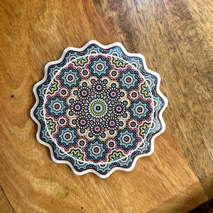 Op de afbeelding: Een ronde onderzetter met een witte, geschulpte rand. De onderzetter heeft een kleurrijk, ingewikkeld mandala-ontwerp met rode, blauwe, gele en bruine patronen. De onderzetter ligt op een houten oppervlak. Diameter: ongeveer 10 cm.