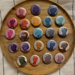May include: A collection of twenty-one round, colorful pinback buttons arranged on a wooden tray. Each button features a unique illustrated character portrait with a name printed above the image. The buttons vary in color, with names like "Sarge", "Doc", and "Caboose".