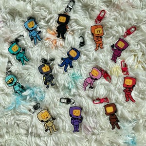 May include: A selection of robot-themed keyrings in various colours. Each keyring features a unique robot design in shades of blue, orange, purple, and yellow. The keyrings are attached to silver metal clasps and are displayed on a fluffy white surface.