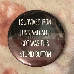 Puede incluir: Un botón redondo con el texto blanco "I SURVIVED IRON LUNG AND ALL I GOT WAS THIS STUPID BUTTON". La mitad superior es negra con efecto de estrellas, y la mitad inferior es de color marrón oscuro.