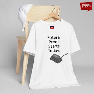 May include: White t-shirt with the text "Future Proof Starts Today." and a graphic of a computer keyboard's Enter key. The shirt is hanging on a wooden hanger, with a white baseball cap resting on a wooden chair.
