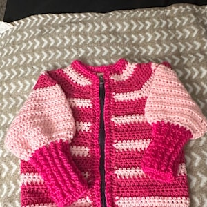 May include: A hand-crocheted jacket in pink and white. The jacket has a full zip, striped design, and a round neckline. The sleeves are a lighter pink, and the cuffs are a darker pink. The jacket is made of soft yarn. The measurements are in centimetres.
