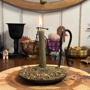 Puede incluir: Una vela verde encendida con una superficie texturizada se encuentra en un soporte de metal oscuro lleno de hierbas secas. La vela está sobre una superficie de madera con un diseño circular. Otros elementos incluyen un cáliz negro y un cuenco de cobre.