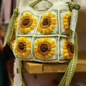 May include: A white crochet handbag with a sunflower design. The bag features yellow and brown sunflowers on a green and white grid pattern. The strap is green and white, and the bag has a handle.