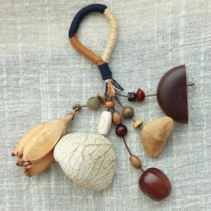 Könnte beinhalten: Ein handgefertigter Schlüsselanhänger mit natürlichen Elementen. Er besteht aus einer Schlaufe aus geflochtenem Kordel in Marineblau, Beige und Braun, verbunden mit einer Kette aus Holzperlen und verschiedenen Samenkapseln. Der Schlüsselanhänger hat eine neutrale Farbpalette.