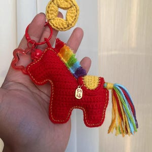Puede incluir: Un llavero de caballo rojo tejido a ganchillo con crin y cola de arcoíris, y un dije dorado. El llavero incluye una moneda tejida a ganchillo amarilla y un cierre rojo en forma de estrella.