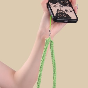 May include: Black phone case featuring a cat design with the text "SWEET CAT". The case is held by a green knitted wrist strap. The phone case has a wooden-like appearance.