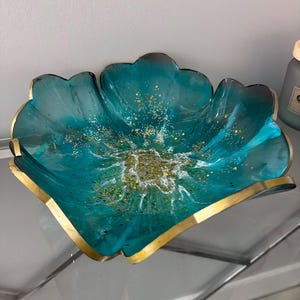 May include: A teal resin bowl with a gold rim and gold glitter accents. The bowl has a flower-like shape with a glossy finish. The interior features white and gold swirls, creating an artistic design. A decorative piece for the home.