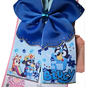 Puede incluir: Un lazo grande azul para el cabello con la palabra "Bluey" y personajes de dibujos animados. El lazo tiene un fondo azul claro con huellas de patas y está adornado con pedrería. El lazo es sostenido por una mano.