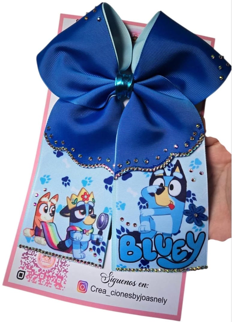 K&ouml;nnte beinhalten: Eine gro&szlig;e blaue Haarschleife mit dem Wort "Bluey" und Zeichentrickfiguren. Die Schleife hat einen hellblauen Hintergrund mit Pfotenabdr&uuml;cken und ist mit Strasssteinen verziert. Die Schleife wird von einer Hand gehalten.