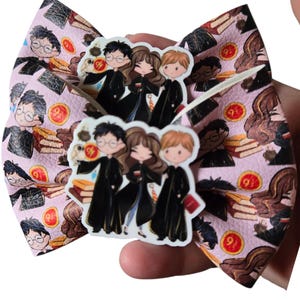 May include: A pink hair bow featuring a repeating pattern of cartoon characters, books, and the number 9 3/4. The bow has a central embellishment with three cartoon figures in black robes. The bow is held in a person's hand.