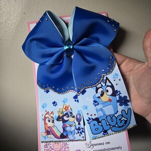 K&ouml;nnte beinhalten: Eine gro&szlig;e blaue Haarschleife mit hellblauem Innenfutter und Strasssteinen. Die Schleife zeigt ein gedrucktes Design von Zeichentrickfiguren und das Wort "Bluey". Die Schleife ist an einer rosa Karte befestigt.