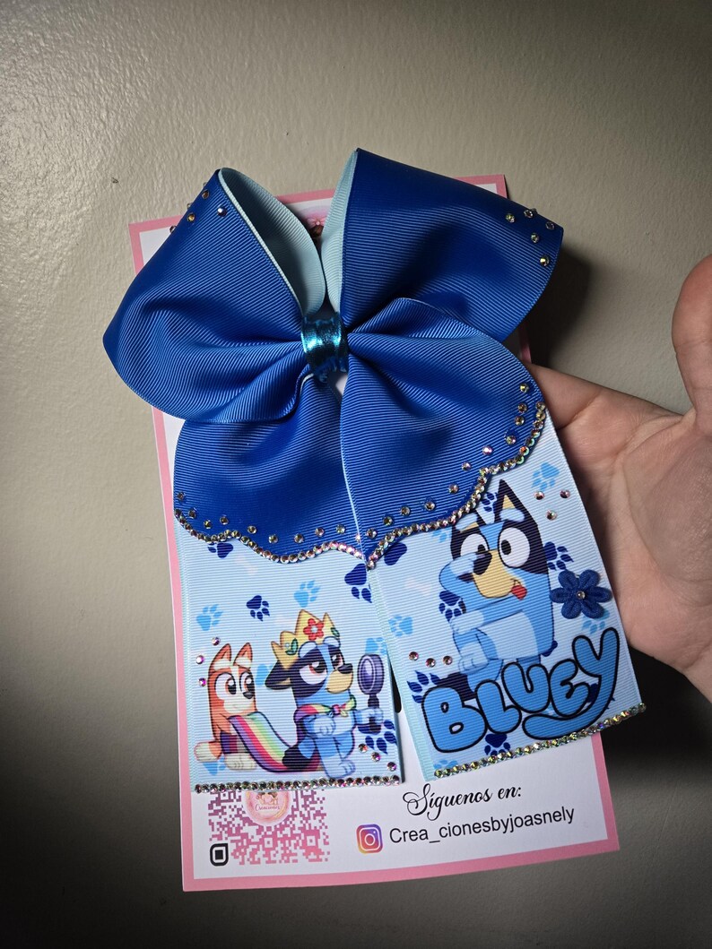 K&ouml;nnte beinhalten: Eine gro&szlig;e blaue Haarschleife mit hellblauem Innenfutter und Strasssteinen. Die Schleife zeigt ein gedrucktes Design von Zeichentrickfiguren und das Wort "Bluey". Die Schleife ist an einer rosa Karte befestigt.