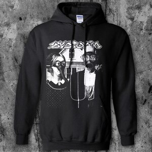 Puede incluir: Sudadera con capucha negra con estampado gr&aacute;fico. El estampado presenta una pareja con m&aacute;scaras de gas frente a una casa, con la palabra "Avaritia" encima en una fuente estilizada. El dise&ntilde;o es en blanco y negro.
