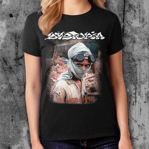K&ouml;nnte beinhalten: Schwarzes T-Shirt mit einem Grafikdruck einer Person, die ein gr&uuml;nes Bandana und eine Schutzbrille tr&auml;gt. Der Text "Dystopia" ist in Wei&szlig; &uuml;ber dem Grafikdruck gedruckt. Der Text "The Last Man" ist in Rot unter dem Grafikdruck gedruckt.