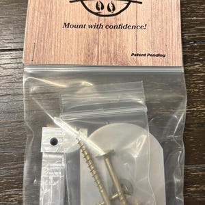 May include: A package containing a mounting kit for hanging items. The packaging features a wooden-textured card with the words "HANG-A-BUCK" and "Mount with confidence!" printed on it. The kit includes screws and metal mounting hardware.