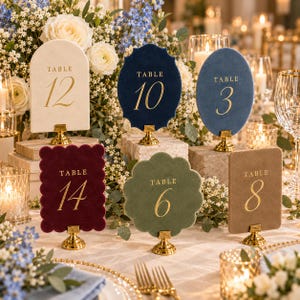 May include: A collection of six velvet table number signs in various shapes and colors, including cream, navy, blue, burgundy, sage green, and tan. Each sign features gold foil text and numbers, supported by small gold metal stands.