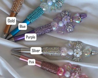 May include: Five decorative pens in gold, blue, purple, silver, and pink. Each pen features a jeweled barrel and a bow or heart-shaped embellishment. The text "Hearts & bows" is at the bottom.