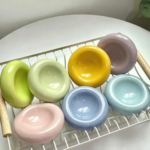 Può includere: Una collezione di sei piccole ciotole in ceramica lisce e circolari in colori pastello, tra cui verde lime, verde chiaro, giallo, lavanda, azzurro e rosa tenue, disposte all'interno di uno scolapiatti in filo metallico bianco con manici in legno.