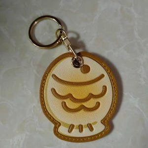 May include: A gold-colored keychain with a fish-shaped charm. The charm features a cream-colored background with a gold outline and gold wave-like designs. The keychain has a gold ring and clasp.