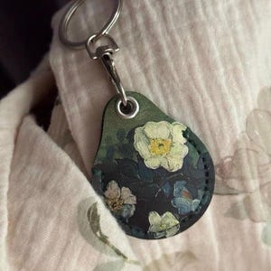 May include: A teardrop-shaped keychain with a floral design. The keychain features a dark green background with white and yellow flowers and blue accents. The metal ring is silver.