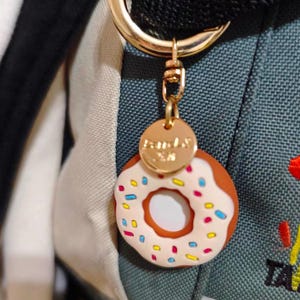 May include: A gold-colored keychain with a donut charm. The donut has a white frosting with colorful sprinkles and a brown base. The keychain is attached to a backpack.