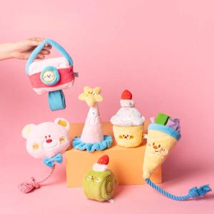 May include: A collection of plush toys in various shapes, including a camera, bear, cupcake, and ice cream cone. The toys feature bright colors like pink, blue, and yellow, with embroidered details and attached rope or ribbon elements.