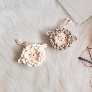 May include: Two handmade crochet sheep ornaments. One is white with pink accents, the other is grey with pink accents. Both have black stitched eyes and a pink nose. Each has a loop for hanging.