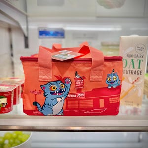 Może przedstawiać: Pomarańczowa, izolowana torba na lunch Trader Joe's z czerwonym zamkiem błyskawicznym i uchwytami. Torba posiada grafikę z kreskówkowym kotem i logo Trader Joe's. Torba znajduje się w lodówce.