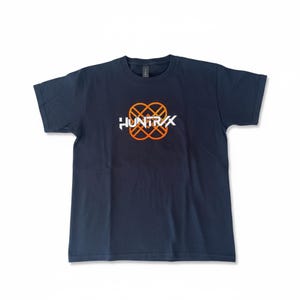 Op de afbeelding: Marineblauw T-shirt met een ronde hals. Het shirt heeft het woord "HUNTRX" in het wit, met een oranje en wit in elkaar grijpend ontwerp boven de tekst. Het shirt is gemaakt van een zacht materiaal.