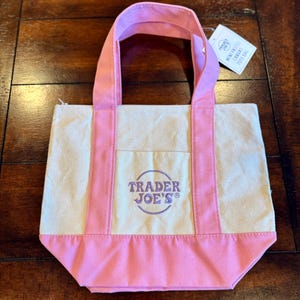 May include: A small canvas tote bag with pink accents. The bag features a cream-colored body with pink handles and a pink bottom. The Trader Joe's logo is printed on the front. A tag reads "Mini Pastel Canvas Tote Bag."