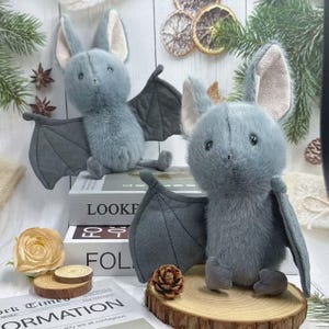May include: Two plush bat toys with gray fur and large gray wings are displayed. The bats have large ears with a light pink interior. The bats are sitting on a wooden surface with a book and a rose in the background.
