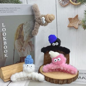 Puede incluir: Cuatro pinzas para el cabello caprichosas con forma de criaturas peludas, cada una con ojos saltones y un gorro de punto. Los colores incluyen marrón, negro, blanco y rosa, con gorros azules y blancos. Una pinza está sujeta a una revista.