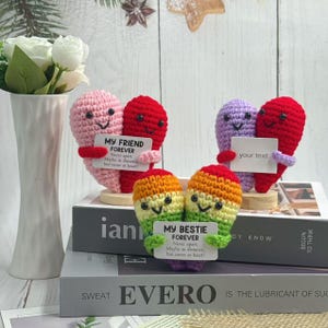 May include: Crocheted heart-shaped figures in pink, red, and purple, each holding a sign. One says "MY FRIEND FOREVER," another "your text," and the third "MY BESTIE FOREVER." The figures are on wooden bases.