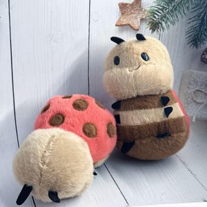 May include: A collection of plush insect toys, including a ladybug and a bee. The ladybug is pink with brown spots, and the bee has brown and tan stripes. The toys are made of soft, fuzzy material and have black antennae.